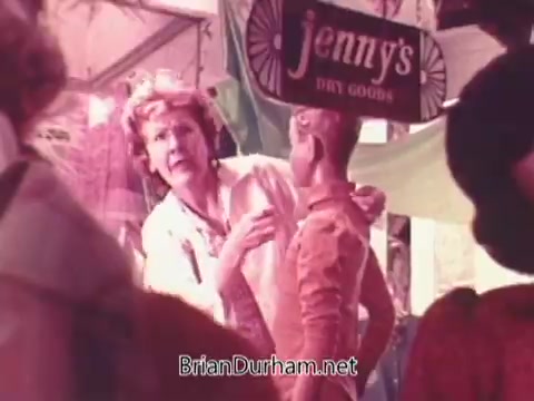 The still from the Oxydol detergent commercial features a woman interacting with a mannequin in a colorful marketplace setting. She appears to be addressing the mannequin with enthusiasm, likely emphasizing the effectiveness of the product. The background includes a sign for 'Jenny's Dry Goods,' contributing to the bustling, vintage shopping atmosphere typical of 1970s commercials.