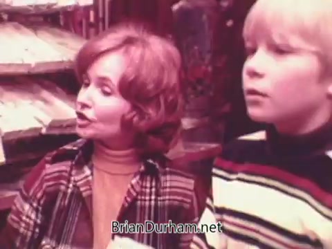 The still from the 1970s Oxydol detergent commercial features a woman with curly hair, wearing a colorful striped outfit, speaking animatedly. Beside her, a young boy in a striped shirt looks on attentively. The background suggests a grocery store setting, indicating a promotional context for the product. The overall vibe is warm and engaging, characteristic of advertising from that era.