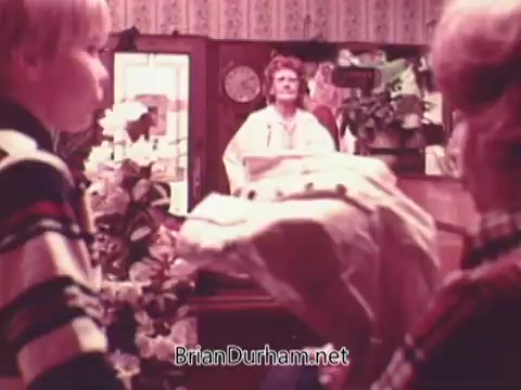 The still from the 1970s Oxydol detergent commercial features a scene with two young boys in striped shirts. They are interacting with a woman at a counter, who is likely a store clerk or a mother figure. The background is decorated with floral arrangements and patterned wallpaper, typical of the era's aesthetic. The mood appears light and engaging, likely emphasizing the effectiveness of Oxydol detergent in a family-oriented context.