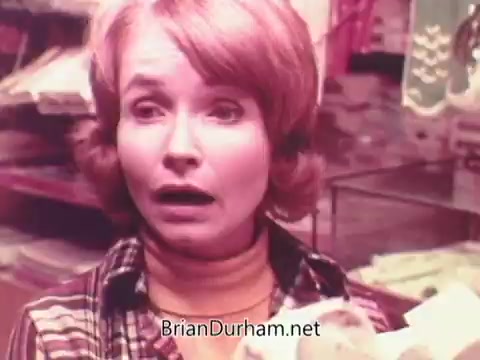 The still from the 1970s Oxydol detergent commercial features a woman with a shocked or worried expression. She has blonde hair styled in soft waves and is wearing a checkered shirt over a turtleneck. The background suggests a home or store environment, likely showcasing laundry or household items. The color palette appears warm, typical of the era's advertising style.