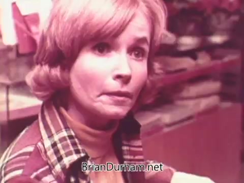 The still from the 1970s Oxydol detergent commercial features a woman with short, wavy hair and a concerned expression. She is wearing a plaid jacket and appears to be in a domestic setting, possibly a kitchen. The colors are warm, contributing to a nostalgic atmosphere typical of that era's advertisements. The context suggests a focus on household care and the effectiveness of the detergent.