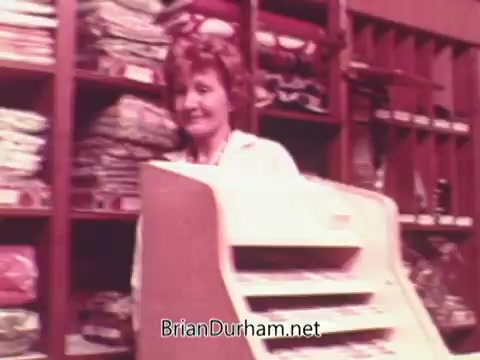 The still from the 1970s Oxydol detergent commercial features a smiling woman standing behind a register in a shop filled with colorful household items. The setting appears dated, with a warm color palette typical of that era. The woman conveys a friendly and inviting demeanor, promoting a sense of reliability associated with the product.