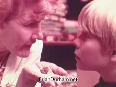 The still from the 1970s Oxydol detergent commercial features a close-up interaction between an older woman and a young boy. The woman appears to be engaging the boy with a serious expression, holding something in her hand as they share a moment of intense focus. The background hints at a domestic setting, possibly a laundry area. The color palette is warm, typical of the era's advertising style. Overall, the scene conveys a sense of connection and attention to detail in the context of laundry care.