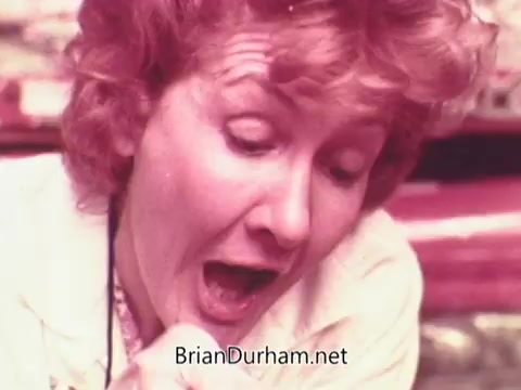 The still from the 1970s Oxydol detergent commercial features a woman with light-colored, curly hair, displaying an exaggerated expression of surprise or excitement. She appears to be engaged in a home-related activity, likely demonstrating the effectiveness of the detergent. The vibrant colors and lively ambiance reflect the advertising style of that era.