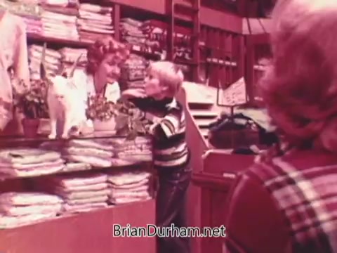 The still from the 1970s 'Oxydol detergent' commercial features a cozy laundry scene. A smiling woman at a counter interacts with a young boy, who is dressed in a striped shirt, while a cat sits nearby. The setting is warm and cluttered, with neatly folded laundry in the background and potted plants, conveying a friendly, nostalgic atmosphere typical of home-care products of that era.