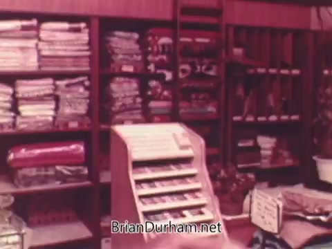 The still depicts a vintage store interior filled with neatly stacked fabrics and various household items on shelves. Prominently featured is a display rack, likely intended for retail sales, showcasing merchandise in an inviting manner typical of 1970s decor. The warm colors and organized layout contribute to a nostalgic atmosphere representative of that era's shopping experience.