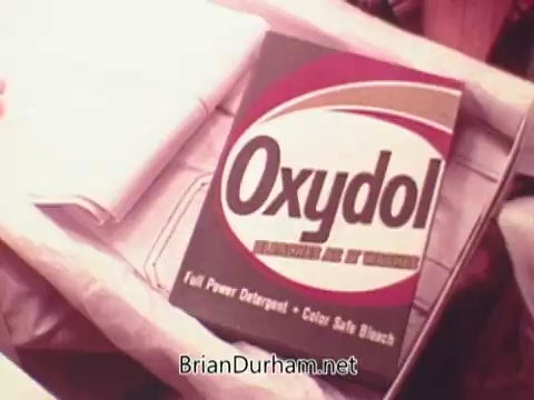 The still depicts a box of Oxydol detergent, prominently featuring its logo against a simple background. The packaging highlights that it is a 'Full Power Detergent' and 'Color Safe Bleach.' The setting suggests a domestic environment, possibly emphasizing the product’s use for laundry. The overall aesthetic has a vintage 1970s feel, characteristic of that era's advertising style.