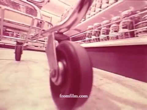 The still from 'Chatham Supermarket Commercials' features a close-up shot of a shopping cart wheel on a retail floor, emphasizing movement through the supermarket. The background blurredly showcases shelves stocked with jars, indicating a focus on products. The aesthetic aligns with 16mm film characteristics, hinting at a nostalgic portrayal of grocery shopping. The associated themes include freshness and variety, with titles like apples, bananas, meats, and steak suggesting a diverse array of grocery offerings.
