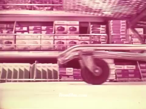 The still from the 'Chatham Supermarket Commercials' features a close-up of a shopping cart wheel rolling across the floor, set against the backdrop of neatly stacked grocery products in a supermarket. The muted colors give it a vintage feel, characteristic of 16mm prints. The scene conveys a lively shopping atmosphere, showcasing various grocery items like apples, bananas, and meats in a prominent supermarket. The focus is on the shopping experience and the selection available at Chatham.