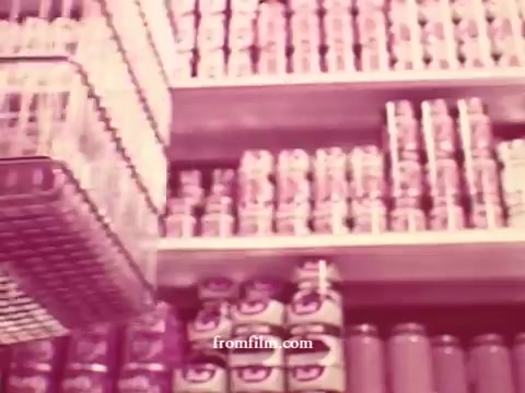 The still features a retro supermarket scene, showcasing shelves filled with canned goods and products typical of a grocery store setting. There's a shopping cart visible, suggesting a focus on shopping convenience. The warm color tones of the 16mm print enhance the nostalgic vibe, reflecting a time when such commercials emphasized fresh produce like apples and bananas, alongside meats and potatoes. The overall aesthetic captures the essence of classic supermarket advertising.