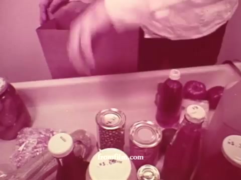 The still from 'Chatham Supermarket Commercials' features a hand placing items into a paper bag. Various grocery products are visible on a conveyor or surface, including jars, cans, and bottles, suggesting a focus on shopping and product variety. The color tone is likely warm or muted, typical of 16mm prints, emphasizing a nostalgic or vintage commercial style. The context hints at promoting fresh produce, meats, and coupons.