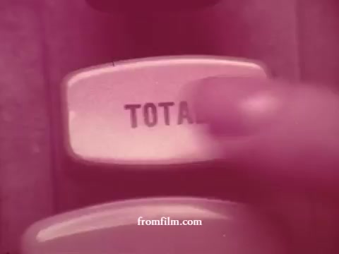 The still from the 'Chatham Supermarket Commercials' features a close-up of a finger pressing a button labeled 'TOTAL' on a vintage cash register or calculator. The visual style is nostalgic, likely reflecting the era of the original 16mm print. The focus on the button suggests an important moment in the checkout process, emphasizing the commercial theme of grocery shopping and total costs, related to items such as apples, bananas, and meats.