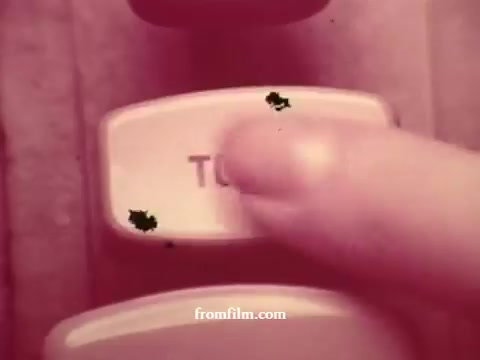 The still from 'Chatham Supermarket Commercials' features a close-up of a hand pressing a button labeled 'TWO.' The image, derived from 16mm prints, exhibits vintage film characteristics, with a slight reddish hue and some visual artifacts. This moment may signal a transition or selection related to various supermarket offerings like apples, bananas, and meats, aligning with the commercial's focus on fresh groceries and savings.