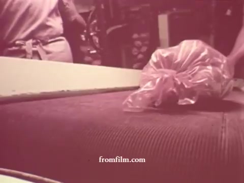 The still from the 'Chatham Supermarket Commercials' captures a behind-the-scenes moment in a grocery setting. A bag of fresh meat is prominently featured, likely showcasing the supermarket's dedication to quality. The vintage aesthetic, characteristic of 16mm prints, offers a nostalgic glimpse into the supermarket environment, while subtle details hint at the variety of products available, including fruits like apples and bananas, and staples like potatoes and steaks. Overall, it reflects the everyday shopping experience of that era.