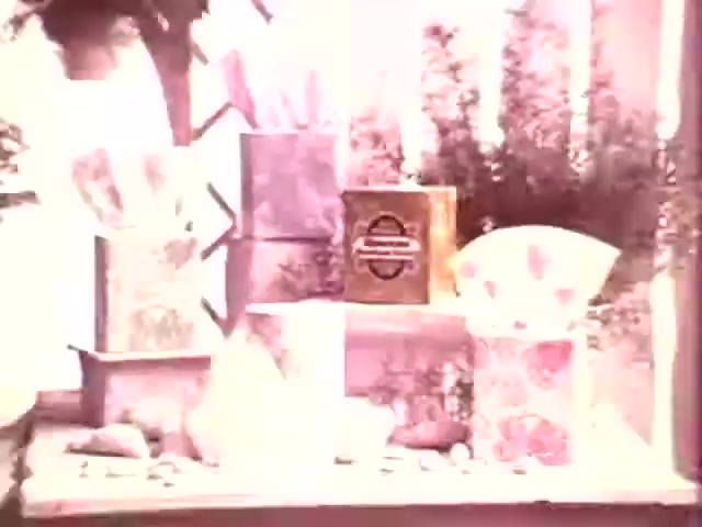 The 16mm still features an arrangement of Kleenex Boutique Tissues boxes, showcasing various vintage designs and pastel colors typical of the 1970s aesthetic. The composition includes multiple tissue boxes stacked and displayed among decorative elements like stones and greenery, reflecting a charming and elegant presentation suited for home décor.