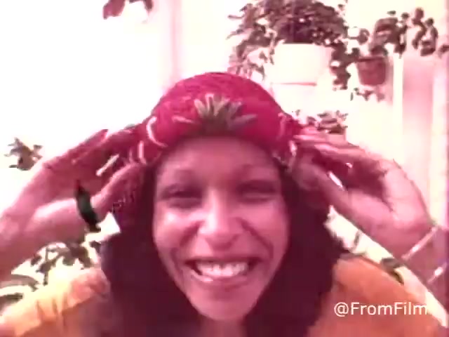 The still from the 1970s 'Kleenex Boutique Tissues' shows a smiling woman wearing a colorful hat. She has long hair and is playfully adjusting the hat while surrounded by verdant plants in a bright setting. This image likely reflects the playful and vibrant marketing style of that era for the Kleenex Boutique Tissues.