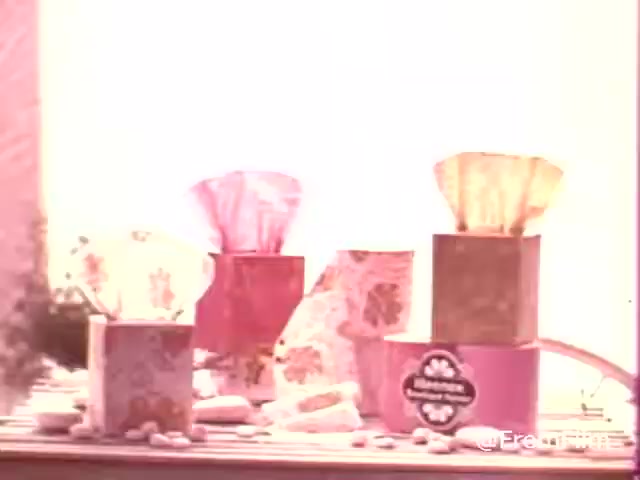 The still features a collection of vintage Kleenex Boutique Tissues boxes from the 1970s. The boxes are decorated with floral patterns and come in various colors, including pink, gold, and other pastel shades. They are arranged on a surface, possibly a table, surrounded by small decorative items. The overall aesthetic is nostalgic, reflecting the design trends of the era. The lighting is soft and warm, enhancing the vintage feel.