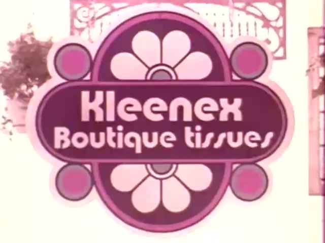 The still features a vibrant and stylized logo for 'Kleenex Boutique Tissues,' characteristic of the 1970s design aesthetic. The logo incorporates bold colors, including purple and white, with decorative floral elements. This visual suggests a playful yet sophisticated product aimed at the boutique market, reflecting contemporary trends of that era. The image is likely derived from a 16mm print, giving it a vintage quality.