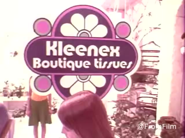 The still features a colorful, retro advertisement for 'Kleenex Boutique Tissues,' prominently displayed with a 1970s aesthetic. The sign has bold text and decorative elements, surrounded by a soft, pastel-colored environment. A person in the background, dressed in a vibrant outfit typical of the era, adds to the nostalgic feel of the scene. The overall composition evokes a sense of playful charm associated with that time period.