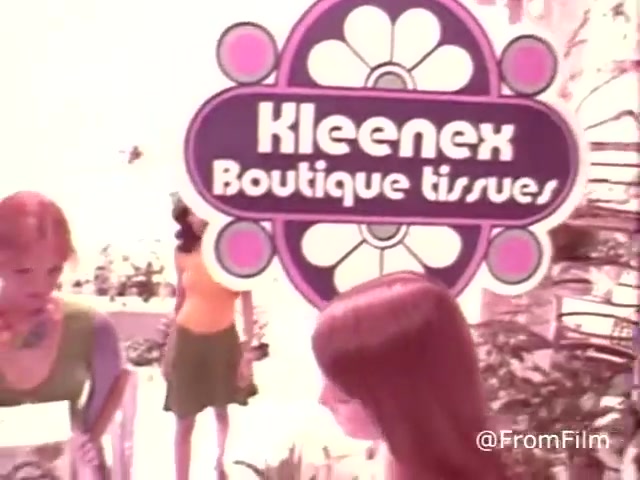 The still from 'Kleenex Boutique Tissues - 1970's' features a vibrant, retro setting with a prominent sign displaying the product name 'Kleenex Boutique Tissues.' The sign showcases a playful design typical of the 1970s aesthetic. In the foreground, two women are engaged with the product, wearing colorful outfits that reflect the fashion of the time. The background includes lush greenery, enhancing the cheerful and lively atmosphere of the scene. The overall look emphasizes a fun and stylish approach to everyday items.