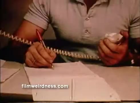 The still features a close-up of a man’s hands engaged in a task related to the Helgoland fisheries research sponsorship. He is holding a telephone receiver with one hand while using a pen to write on a sheet of paper with the other. His shirt is casual, suggesting an informal setting. The focus conveys a sense of organization and communication, likely tied to the promotion of fisheries research initiatives.