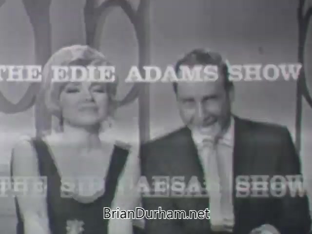 The still features Edie Adams and Sid Caesar smiling at the camera, likely promoting their show from 1963. The backdrop consists of a stylized design, and the text overlay prominently displays 'THE EDIE ADAMS SHOW' and 'THE SID CAESAR SHOW.' The image captures a lighthearted, engaging moment between the two, reflecting the entertainment style of the era.