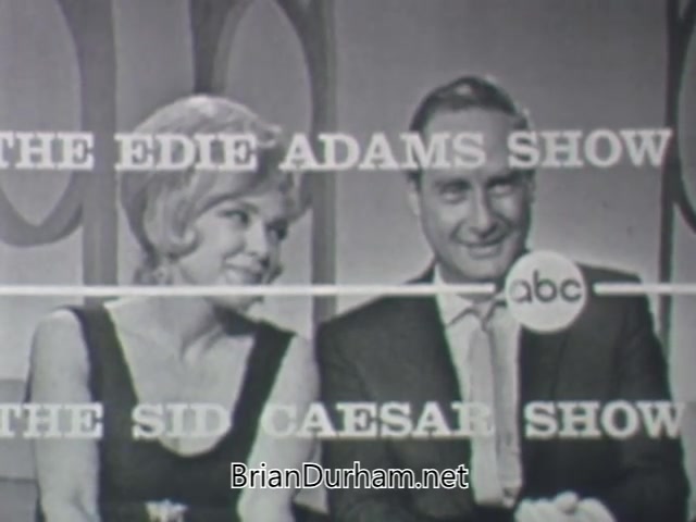The still features Edie Adams and Sid Caesar seated side by side, promoting their show in 1963. Edie wears a black dress and has styled blonde hair, while Sid is in a suit and looks engaged. The background includes circular shapes and text that reads 'THE EDIE ADAMS SHOW' and 'THE SID CAESAR SHOW,' indicating a television promo for their collaboration on ABC. The overall vibe is retro and captures the essence of early 1960s variety shows.