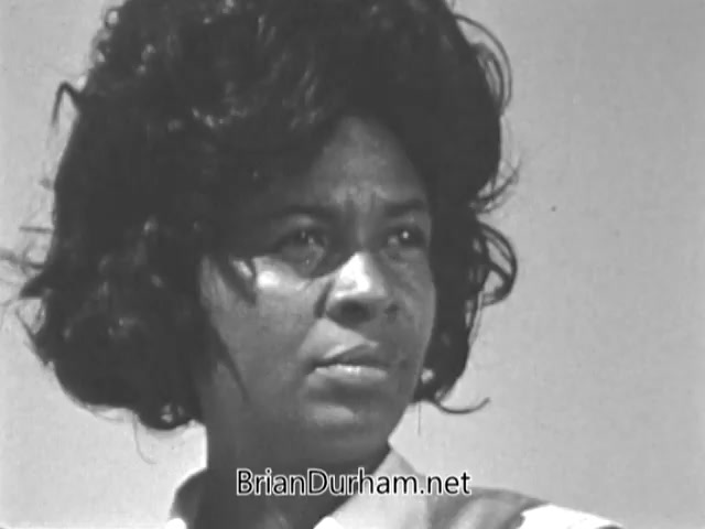 The still features a close-up portrait of a woman with a thoughtful expression, emphasizing her determination and focus. She has voluminous, curly hair, and is wearing a light-colored collar, suggesting a context of service or community engagement. The background is bright and minimally detailed, drawing attention to her as a representative of the VISTA program and its mission to combat poverty through volunteerism.