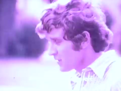 The still from the Gaines Burgers dog food commercial features a young man with curly hair, possibly in profile, set against a softly blurred background. The color palette is muted, leaning towards a light purple hue that gives the image a nostalgic feel, characteristic of the 1970s era. This aesthetic aligns with the playful and family-oriented marketing of Gaines Burgers during its production years, highlighting the bond between pets and their owners.
