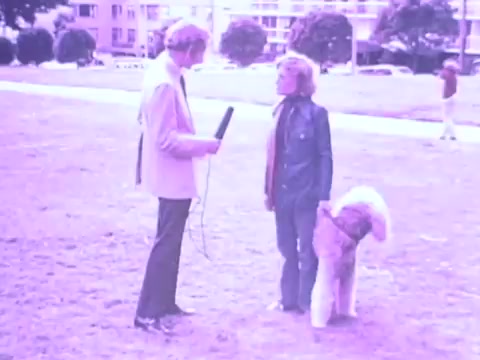 The still from the 1970s Gaines Burgers dog food commercial features a man interviewing a woman in a park, both dressed in typical attire of the era. The scene appears playful, highlighting the connection between dogs and their owners. A fluffy dog stands beside the woman, adding to the lighthearted atmosphere. The image has a distinctive purple tint, characteristic of the film's aesthetic from that time. This captures the nostalgic essence of dog ownership and the promotion of Gaines Burgers.
