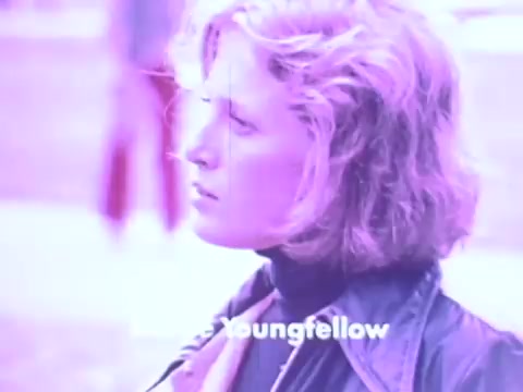 The still from the Gaines Burgers dog food commercial features a woman with shoulder-length hair, appearing contemplative. She is dressed in a leather jacket and turtleneck, suggesting a stylish, contemporary look for the 1970s. The image has a purplish tint, which might be a result of the film's processing or color grading. The commercial likely emphasizes a connection between quality dog food and pet care, reflecting the era's marketing approaches.