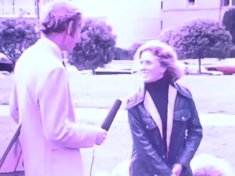 The still from the Gaines Burgers dog food commercial features a male presenter and a female interviewee in a park-like setting. The woman, smiling, appears to be discussing something related to dogs, possibly the product. The male presenter, dressed in a light suit, is holding a microphone, suggesting that they are filming an interview. The color tone of the image has a vintage, slightly faded look typical of 1970s film, enhancing the nostalgic feel of the era. In the background, trees and a car are visible, contributing to the casual outdoor atmosphere.