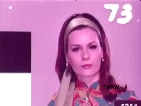 The still features a woman with a stylish appearance, wearing a colorful patterned outfit and a headband. She has a neutral expression and is set against a soft pink background. The image has a vintage quality, characteristic of a 16mm film, and includes a timestamp in the corner. This context suggests it is part of an advertisement for Flair Pens, aiming to convey elegance and style.