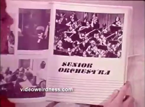 The still features a person holding an open book or magazine page with images showcasing a 'Senior Orchestra.' One prominent photo depicts musicians playing in an orchestral setting, likely highlighting the importance of music and community engagement in promoting better hearing. The layout includes bold text labeling the section 'SENIOR ORCHESTRA.'