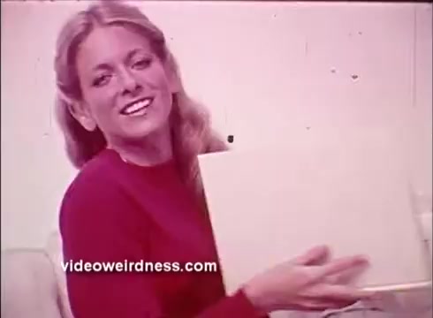 The still from the PSA for the Better Hearing Institute features a smiling woman wearing a red sweater, sitting and holding a white box. She appears cheerful and engaging, likely aiming to convey a positive message about hearing health. The background is neutral, enhancing the focus on her expression and the box.