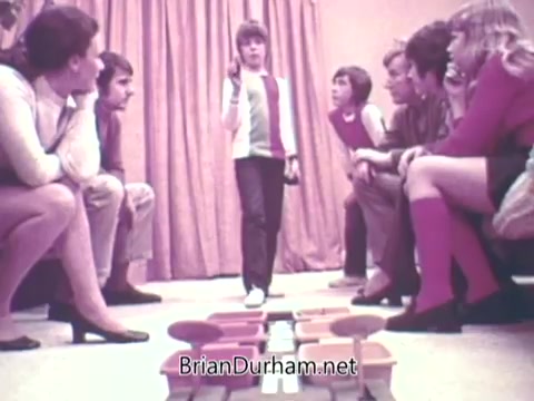The still from the 1973 'Ideal 'Up Set' game' features a group of children seated in two rows facing a central game setup with colorful game pieces. One child stands in the middle, appearing to explain or demonstrate the game. The setting includes a curtain in the background, emphasizing a playful and casual atmosphere. The group's attire reflects the style of the early '70s, with some wearing vests and knee-length socks.