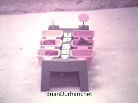 The still features a vintage board game titled 'Up Set,' produced by Ideal in 1973. The game consists of a colorful arrangement of plastic containers, some of which are filled with items, with a black base displaying the game's title prominently. The background appears blurred, highlighting the game's structure.