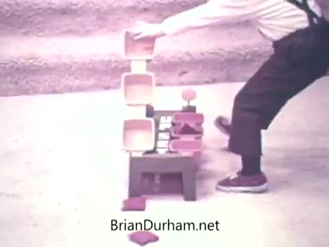The still depicts a scene from the 1973 'Ideal Up Set' game, featuring a person manipulating colorful stacking blocks. The setup includes several shapes stacked on a base, with a focus on the act of rearranging or balancing components. The background is simple, emphasizing the activity. The image conveys a playful, interactive experience typical of children's games from that era.