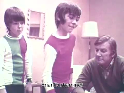 The still from the 1973 'Ideal 'Up Set'' game features two boys and an adult man in a casual, cozy setting. The boys are dressed in colorful sweaters with bold patterns, while the man appears to be engaged in a game or activity with them. The background suggests a home environment, with simple furnishings and a warm atmosphere. The focus seems to be on interaction and playfulness among the characters.