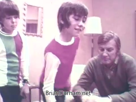 The still from 'Ideal 'Up Set' game - 1973' shows two boys and an adult man in a cozy indoor setting. The boys, dressed in colorful sweaters, appear engaged and animated, while the man, wearing a sweater, seems to be explaining something or participating in a game with them. The atmosphere feels friendly and playful, indicative of a family-oriented game experience. The background includes simple furnishings typical of the era.