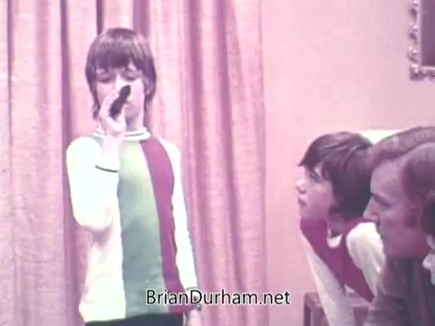 The still features a young boy standing and holding a microphone, singing or speaking. He wears a brightly colored shirt with green, red, and white stripes. Two seated figures, likely an adult and another child, are attentively watching him. The background shows a curtain that adds a warm, homey atmosphere, typical of the early 1970s. The scene captures a moment of family engagement, possibly during a game or entertainment activity.