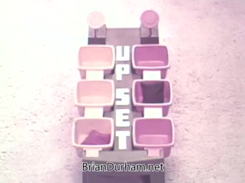 The still features a vintage game setup titled 'UP SET,' with a simplistic design. It consists of several colored containers arranged in two columns, labeled prominently with the word 'UPSET' in bold letters at the center. The color scheme includes various shades of pink and white, indicative of the 1970s style. The background appears neutral, likely intended to emphasize the game elements themselves.