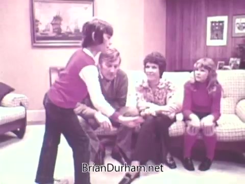 The still from 'Ideal 'Up Set' game - 1973' features a scene in a living room where a young boy is standing and appears to be explaining or demonstrating something, while three adults are seated on a couch watching him attentively. The setting has a warm, 1970s aesthetic, with wood paneling and vintage decorations. The expressions of the seated individuals suggest engagement and interest in what the boy is presenting.