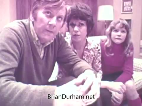 The still from the 1973 'Ideal 'Up Set'' game features a family group of three individuals. A man in a sweater and collared shirt sits in the foreground, looking intently at something off-camera. Next to him, a woman with a bob haircut appears engaged, while a young girl in a red sweater sits beside her, expressing curiosity. The background includes a homey setting with a lamp and patterned upholstery, suggesting a casual family gathering or gameplay atmosphere.