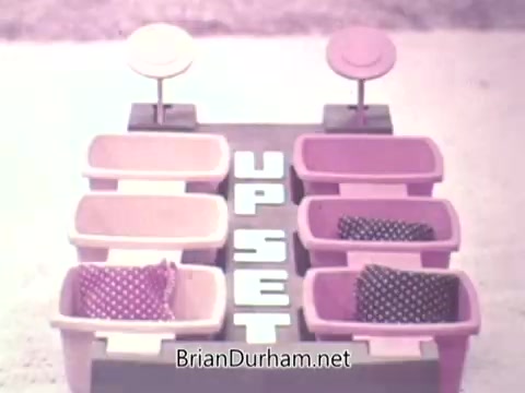 The still from the 1973 'Ideal Up Set' game features a colorful game board with sections labeled 'UP SET.' There are pink and purple bins arranged in two rows, each containing polka-dotted pillows. The overall design is bright and playful, reflecting the style of children's games from that era.