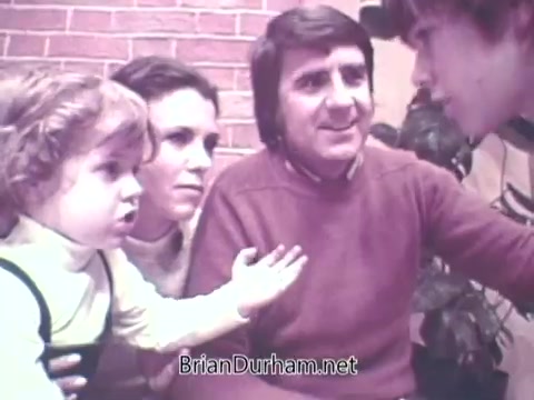 The still from the 1973 'Ideal Up Set' game features a family scene with four individuals. A young girl, dressed in a playful outfit, is animatedly expressing herself, while a woman beside her looks on with concern. A man in a sweater appears engaged in the conversation, and a teenage boy is also involved, seemingly in discussion. The backdrop is a brick wall, suggesting a casual home environment. The overall mood conveys a mix of excitement and tension typical of family game interactions.