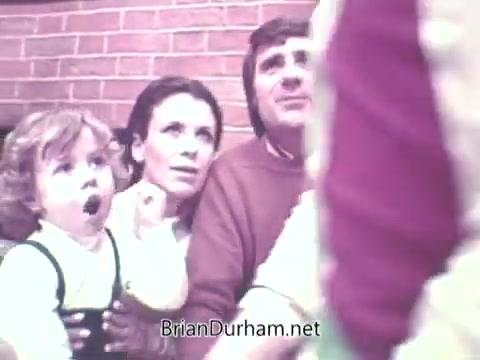 The still features a family scene from the 1973 'Ideal Up Set' game. A young child with curly hair looks surprised or excited, while an adult woman beside them displays a similar expression of interest. Another adult male appears engaged in the moment. The background shows a brick wall, suggesting a cozy, domestic setting. The overall mood is one of anticipation and engagement with the game.