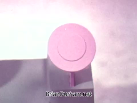 The still from the 16mm film 'Ideal 'Up Set' game - 1973' features a close-up view of a pink circular object, potentially a game piece or component. The background appears to be muted, drawing attention to the pink element. The simplistic design suggests a playful or nostalgic theme, typical of board games from that era.