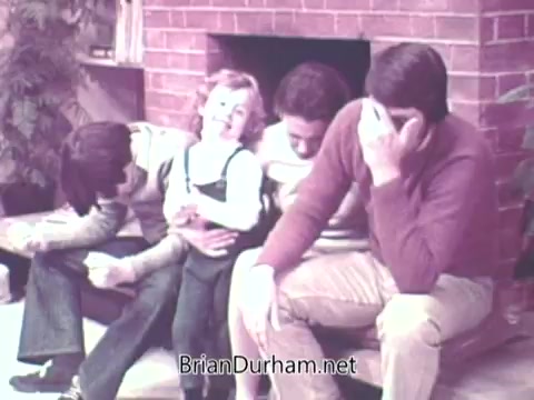 The still from 'Ideal 'Up Set' Game - 1973' features a group of four people sitting together in a cozy interior setting with a brick fireplace. A young child is prominently seated in the center, engaged in the moment. The adults around the child display a range of expressions, with one covering their face, indicating a playful or humorous situation. The overall atmosphere seems casual and warm, likely reflecting a family or friendly gathering.