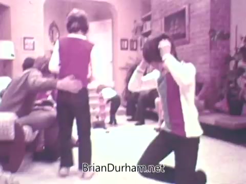 The still from 'Ideal 'Up Set' game - 1973' captures a lively scene in a living room setting. Several children and adults are engaged in play, with one child, wearing a pink and white top, standing with a joyful expression. Another child is kneeling nearby, while a few others are visible in the background, adding to the playful atmosphere. The room features a cozy, homey design typical of the 1970s.